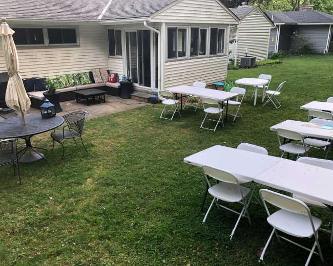 A grassy backyard features several empty folding tables and chairs arranged for an outdoor gathering, with a house visible in the background.