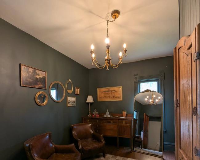 The image depicts a dimly lit, rustic room featuring two brown leather chairs, a wooden sideboard, a mirror, and various framed artworks on the walls.
