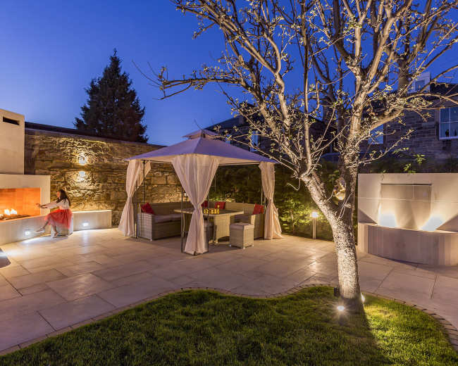A landscaped outdoor area features a seating arrangement under a canopy, a stone fire pit, and a flowering tree.