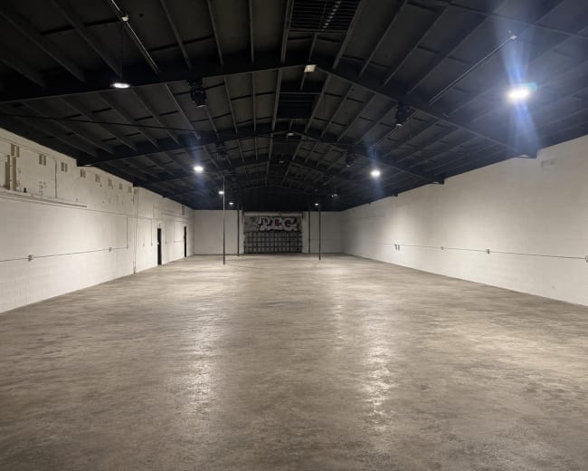 The image shows a large, empty warehouse space with a concrete floor and high, black ceiling, illuminated by overhead lights.