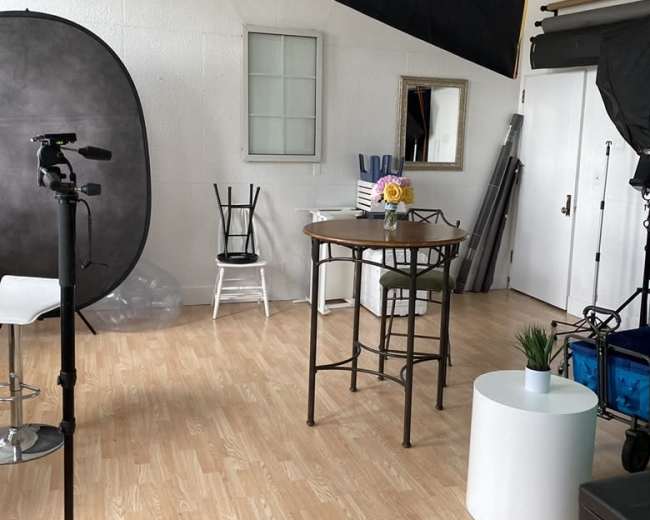 The image shows a photography studio with various equipment, a small round table with a flower vase, and a couple of chairs.