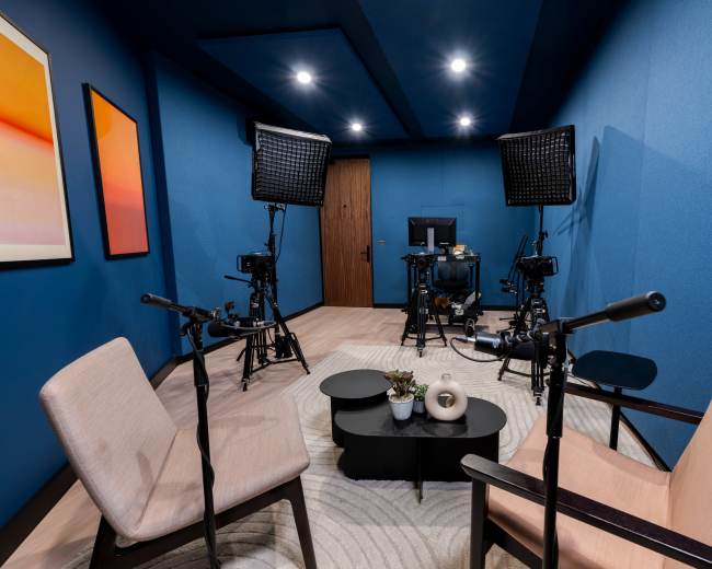 The image shows a small, blue-walled studio equipped with two chairs, a dark coffee table, and several videography lights aimed at a camera setup.