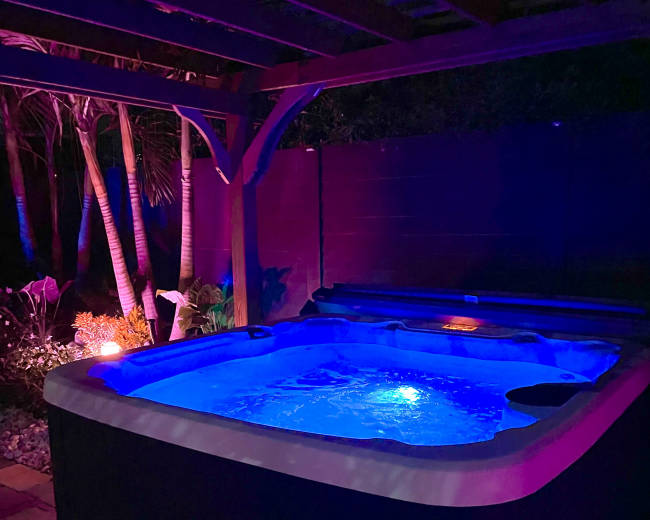 A hot tub illuminated by blue lights sits under a wooden pergola in a garden surrounded by tropical plants.