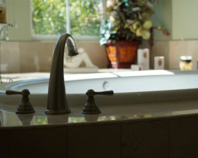 The image shows a bathroom with a large, white bathtub surrounded by light-colored tiles and a window letting in natural light.