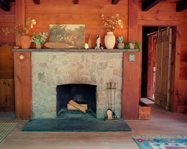 A stone fireplace with a wooden mantel is situated in a wooden-paneled room, surrounded by decorative plants and a painting.