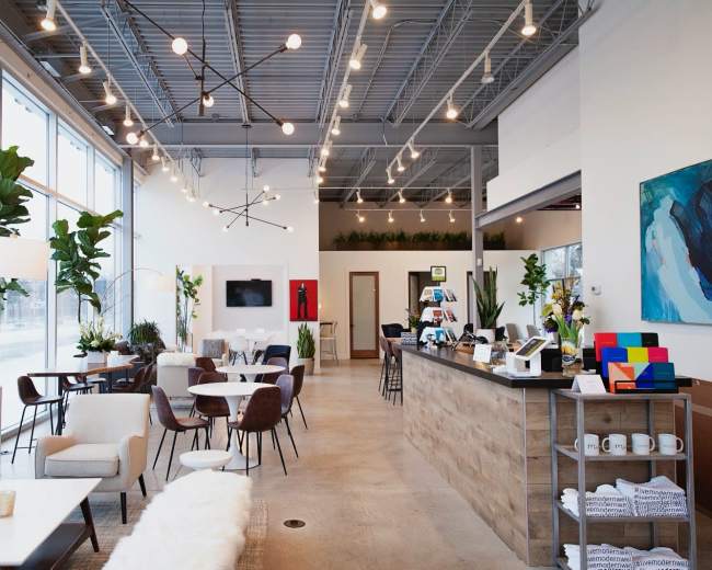 A modern café features a spacious layout with seating areas, a coffee counter, and plants arranged throughout.