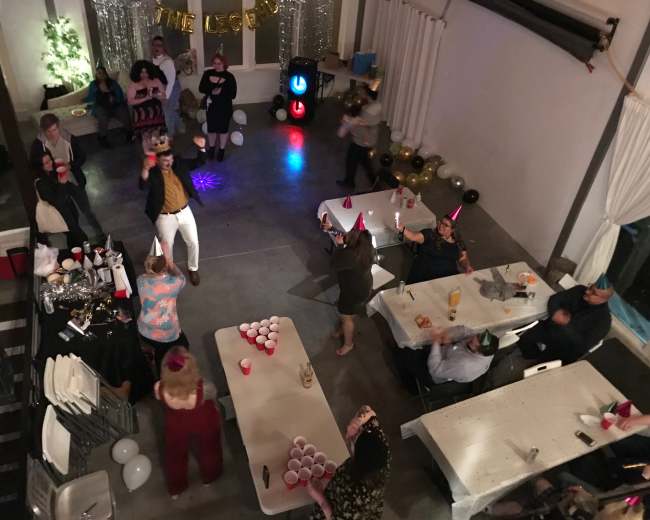 A crowded party scene shows guests dancing and socializing around tables adorned with drinks and decorations, while a colorful light illuminates the floor.
