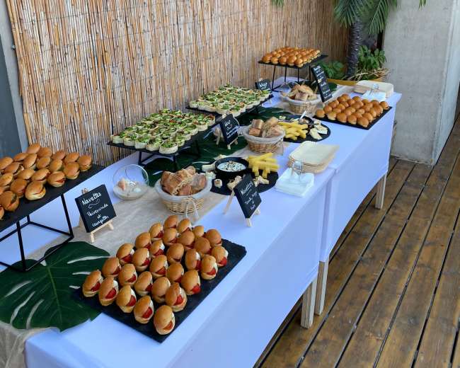 A buffet table is set up with an assortment of snacks, including sliders, desserts, and sushi, against a backdrop of bamboo and greenery.