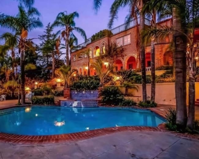 A large swimming pool is surrounded by lush palm trees and a beautifully landscaped yard, with a two-story brick house visible in the background.