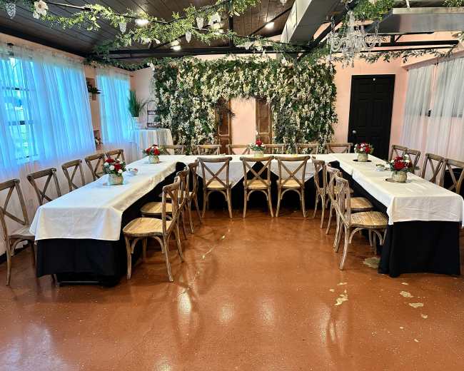 The image shows a decorated event space with two long tables set with white tablecloths, each adorned with floral centerpieces, surrounded by wooden chairs, against a backdrop of artificial greenery on the wall.