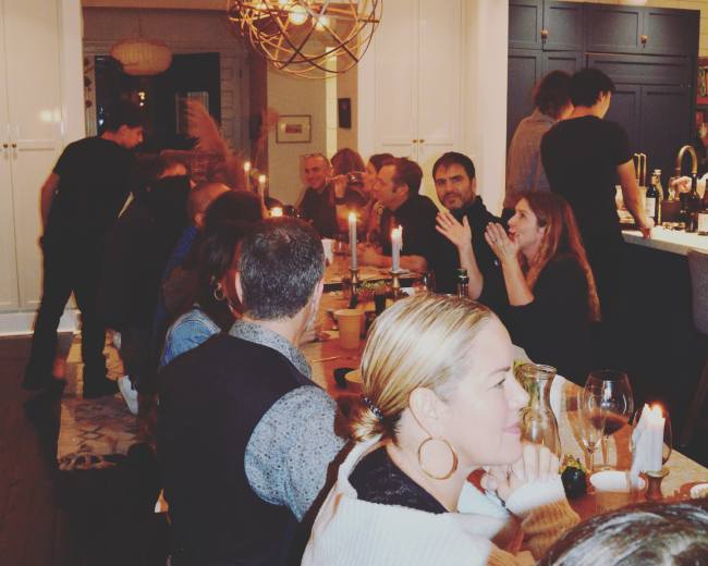 A group of people sit around a long dining table in a well-lit kitchen, sharing a meal with candles and pendant lighting overhead.