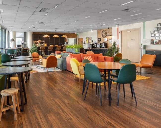 The image shows a spacious modern café with various seating arrangements, including colorful chairs and wooden tables, along with a service area in the background.