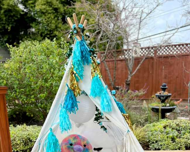 A tent made of white fabric is set up outdoors, decorated with teal and gold accents, and contains plush toys and a patterned mat inside.