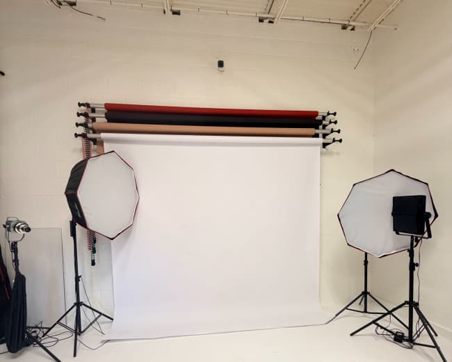 The image shows a photography studio with a white backdrop, two softbox lights on either side, and colored backdrop rolls mounted on the wall.