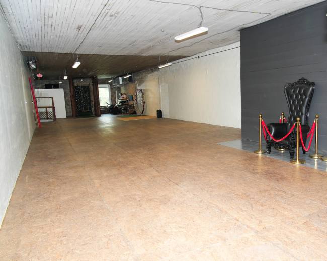 The image shows an empty, spacious room with a wooden floor and a decorative black throne surrounded by red velvet ropes at one end.