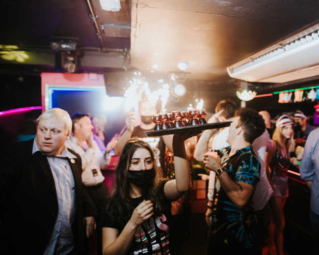 A woman wearing a mask holds a tray of lit sparklers while people celebrate at a crowded bar.