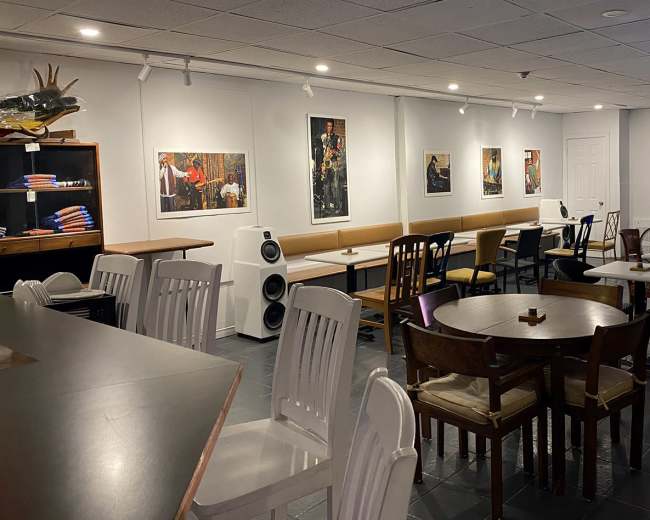 The image shows a spacious café interior with a mix of tables and chairs, featuring artwork on the walls and a counter in the foreground.