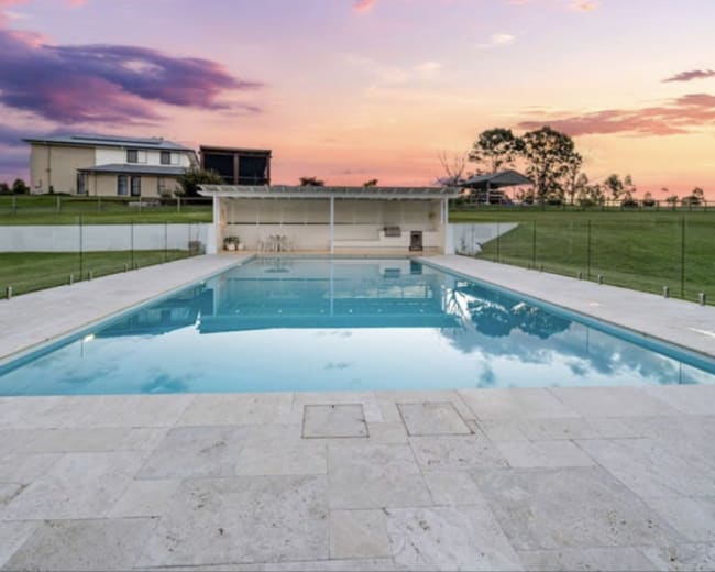 The image shows a rectangular swimming pool surrounded by a stone patio, with a house and trees in the background against a colorful sunset.