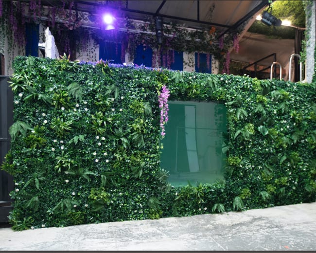 A wall covered in artificial greenery with flowers and a transparent window is shown in an indoor setting.