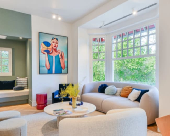The image shows a modern living room featuring a curved sofa, a round coffee table, and a large art piece on the wall, with natural light coming in from large windows.