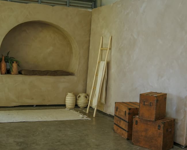 The image shows a minimalist interior space with a curved alcove featuring plants and cushions, a wooden ladder, woven textile on the floor, and stacked wooden boxes.