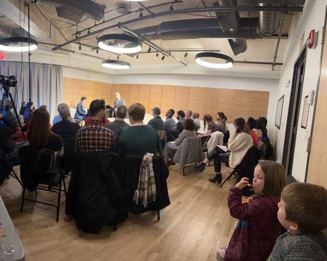 A group of people sits in a semi-circle facing a small stage in a spacious room, with a camera set up for recording.