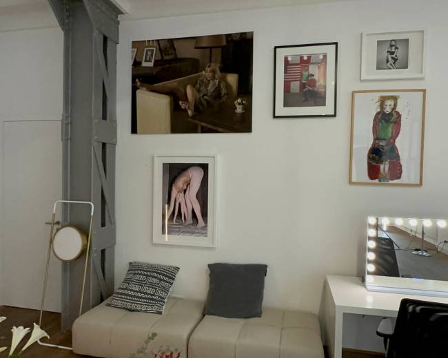 The image shows a room with a white wall adorned with various framed artworks and a mirror with lights, alongside a beige sofa and a desk chair.