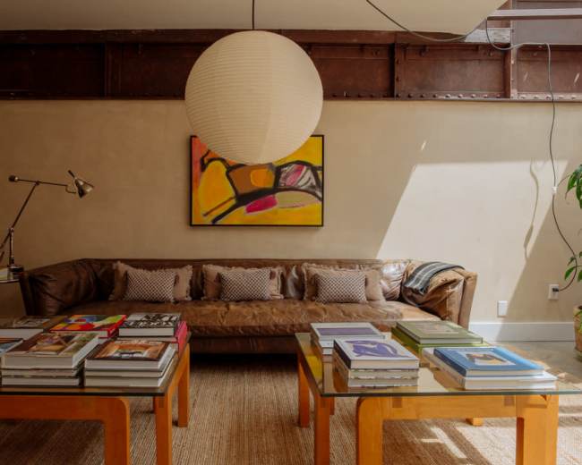 A cozy living room features a brown leather sofa, colorful artwork on the wall, and a coffee table stacked with books.