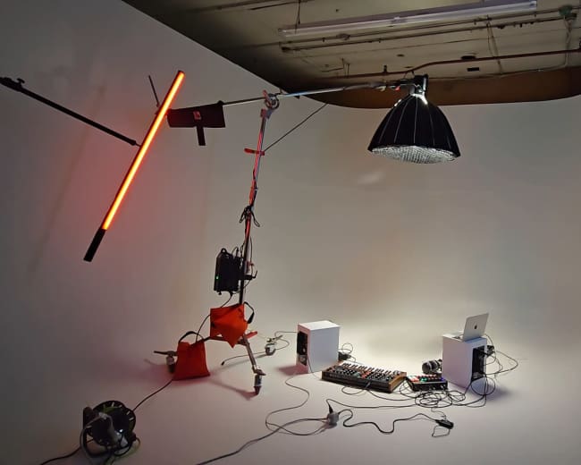 The image shows a minimalistic studio setup with colorful lighting, a music production console, and a laptop arranged against a white background.