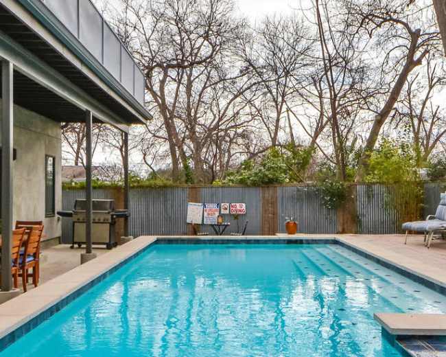 The image depicts a rectangular swimming pool surrounded by a concrete patio, with a backdrop of bare trees and a corrugated metal fence.