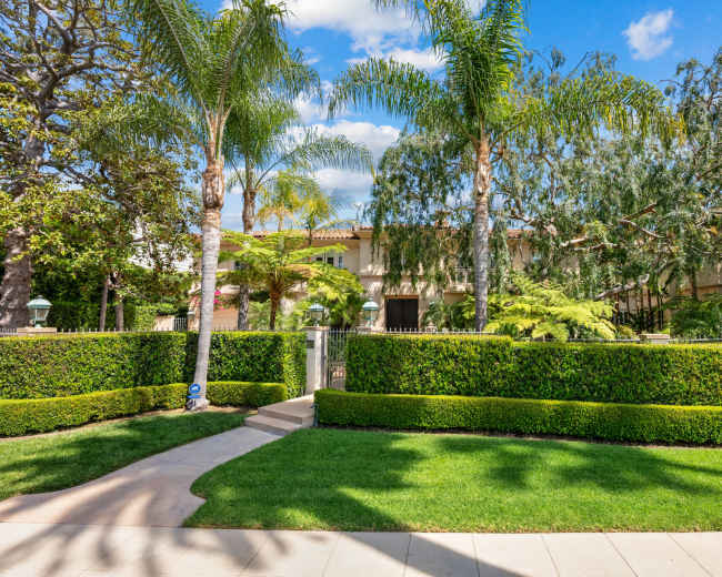 The image shows a landscaped front yard with neatly trimmed hedges, palm trees, and a pathway leading to a large house partially obscured by greenery.
