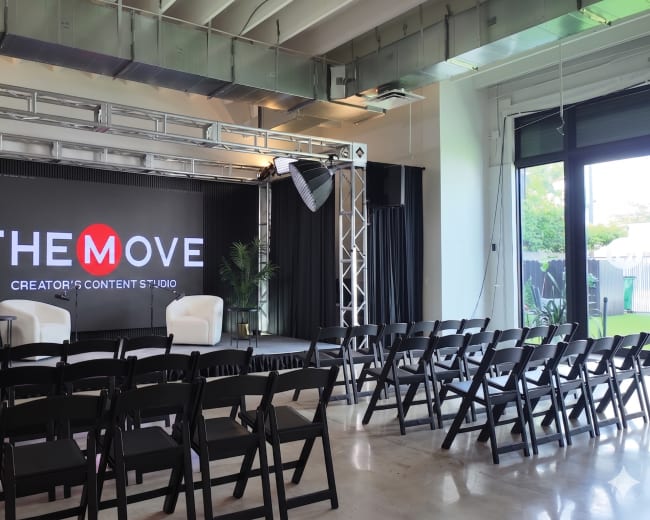 The image shows a modern studio setup with two white chairs on a stage, flanked by black curtains and a large LED screen displaying "THE MOVE CREATOR'S CONTENT STUDIO." Rows of black folding chairs are arranged in front of the stage.