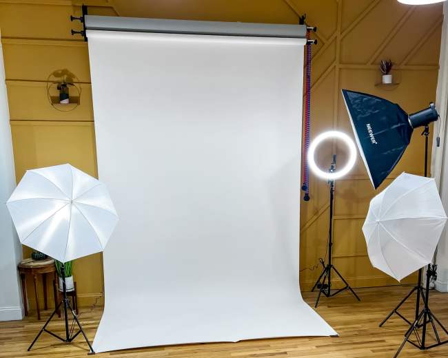 A photography studio setup with a white backdrop, three lighting fixtures, and wooden flooring.