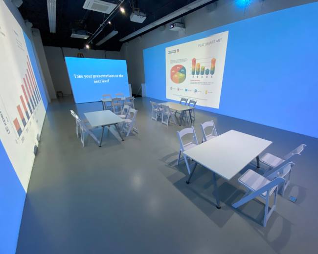 A spacious room with blue walls features several white tables and chairs arranged for a presentation, accompanied by large visual graphics on the wall.