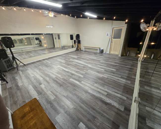 The image shows a spacious dance studio with wooden flooring, a large mirror on one wall, and speaker equipment set up on either side.