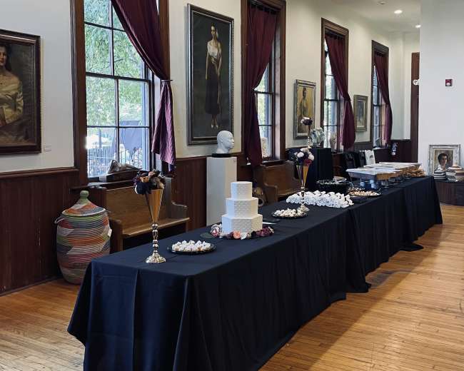 A banquet setup in a room with dark wood accents, featuring tables covered with black cloth and adorned with various decorative items and desserts, along with large portraits hanging on the walls.