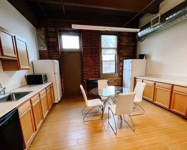 The image shows a kitchen with wooden cabinets, a round glass dining table surrounded by four white chairs, and two refrigerators against a brick wall.