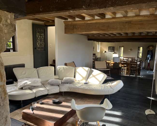 The image shows a modern living space with a curved white sofa, a wooden coffee table, and a dining area in the background featuring wooden furniture.