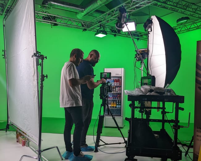 Two individuals are setting up a camera and equipment in a studio with a green screen backdrop.