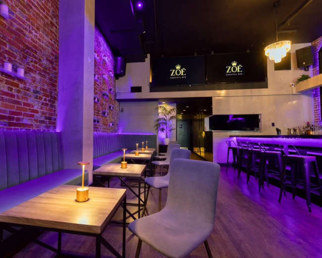 The image shows an upscale bar interior featuring exposed brick walls, purple lighting, and modern furniture with a long seating area and tables.