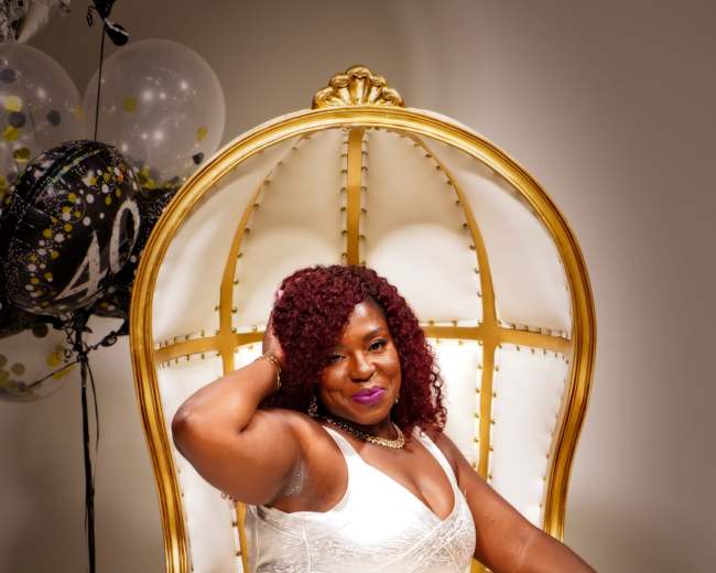 A woman in a white gown sits gracefully in a large, ornate golden chair, surrounded by black and silver balloons celebrating a milestone birthday.