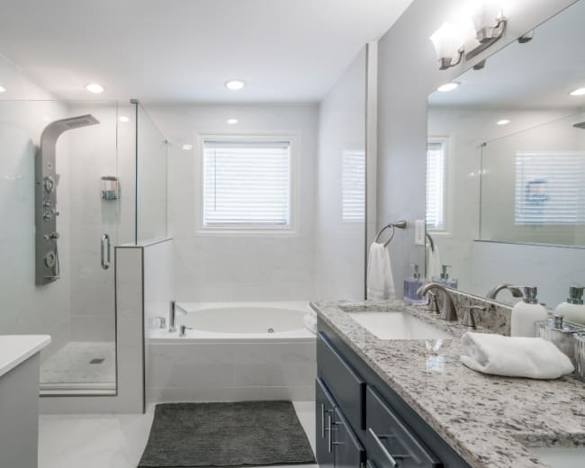 The image shows a modern bathroom featuring a glass shower, a freestanding bathtub, a double sink vanity, and neutral-colored tiles.