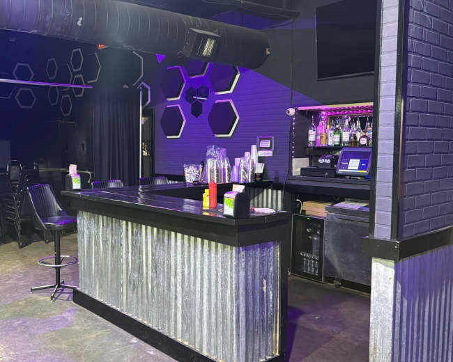 The image shows a dimly lit bar area with a metal counter, shelves stocked with bottles, and hexagonal wall designs.