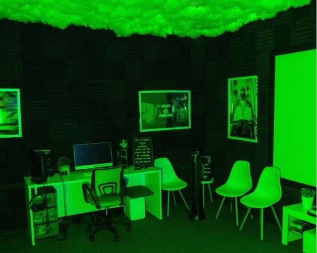 A small room with green lighting, featuring a computer desk, several white chairs, and a large screen with a blank wall behind it.