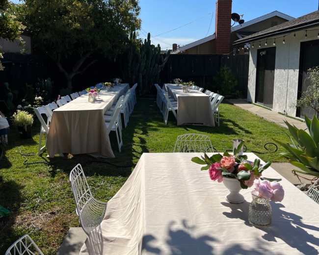 The image shows a well-arranged outdoor setting with tables covered in light fabric, surrounded by greenery and string lights overhead.