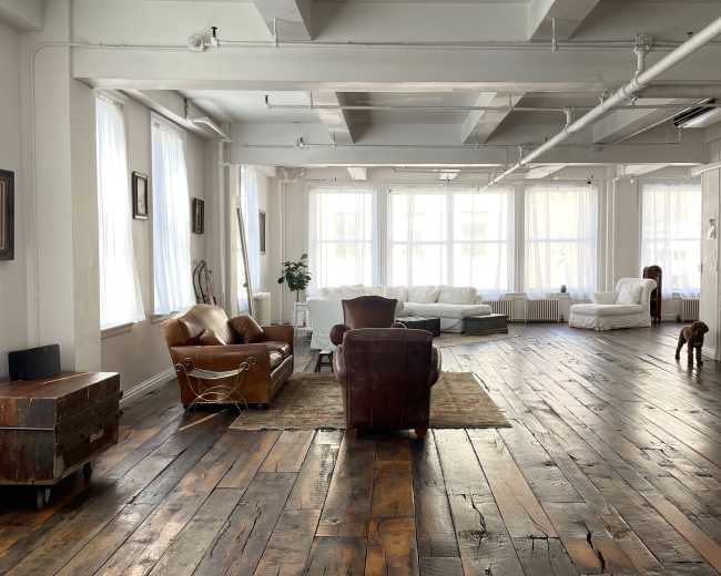 A spacious, bright room with wooden floors, large windows, and a mix of seating areas, including a couch and armchairs.