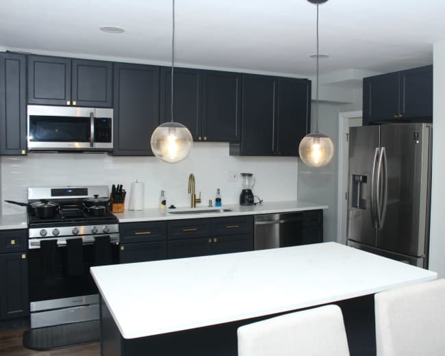 The image shows a modern kitchen featuring dark cabinetry, stainless steel appliances, a large white island with a marble countertop, and pendant lighting.