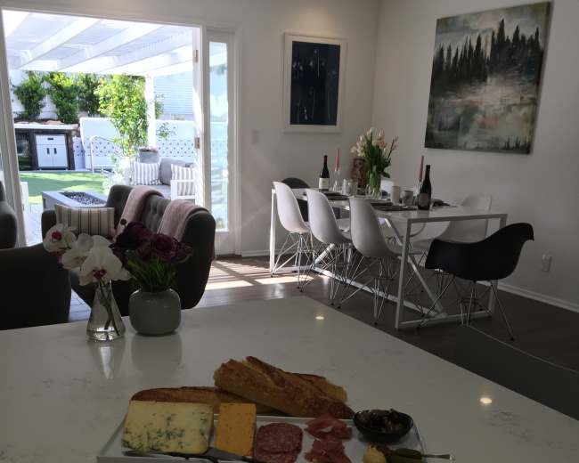 A kitchen counter is set with a charcuterie board, featuring assorted cheeses, meats, and fruits, while a dining area is visible in the background with a table set for a meal.