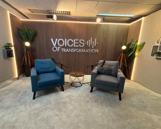 The image shows a modern studio setup with two armchairs, a small table, and a backdrop featuring the text "VOICES OF TRANSFORMATION," illuminated with soft lighting and surrounded by decorative plants.