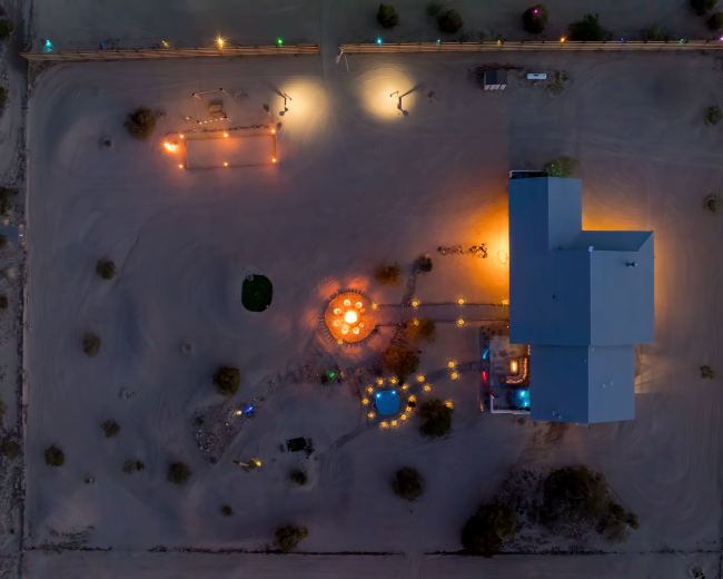 The image shows an overhead view of a large building surrounded by various outdoor features, including lights and a circular arrangement of seating, all set in a sandy area.